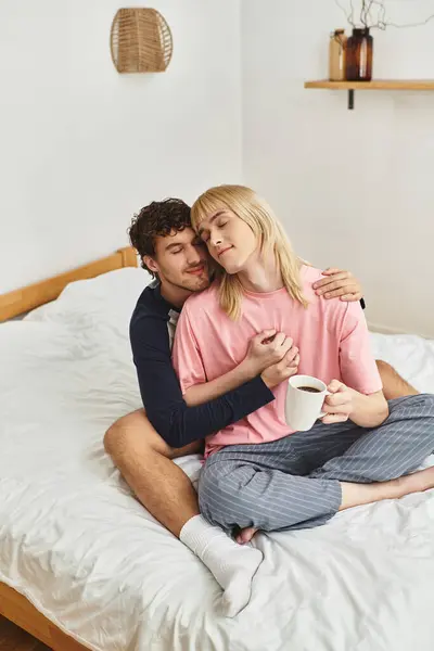 In their bright bedroom, a gay couple enjoys a tender moment while sipping coffee and embracing. — Stock Photo