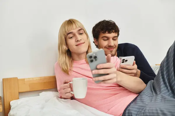 Любящая гей-пара отдыхает в постели, наслаждаясь кофе и занимаясь своими устройствами. — стоковое фото