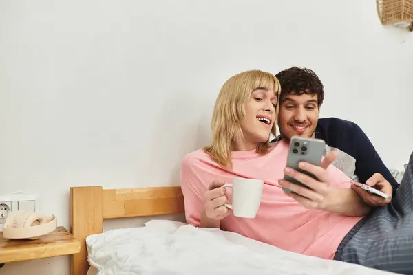Two men cuddle closely on a bed, sharing a joyful moment while looking at a phone, sipping coffee. — Stock Photo