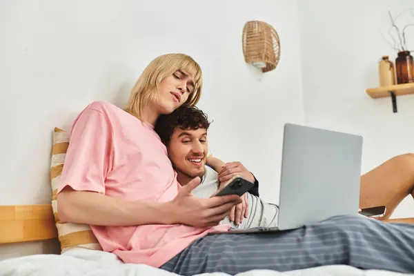 Un couple gay joyeux se détend sur un lit, partageant rires et moments tout en utilisant leurs appareils. — Photo de stock