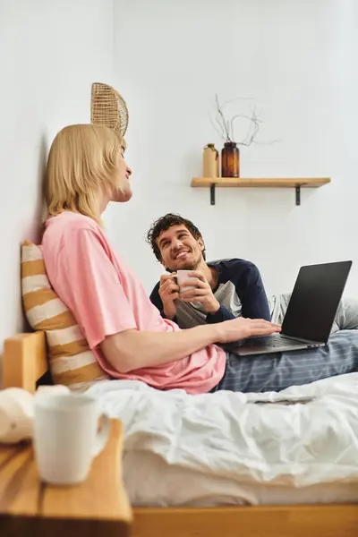 Two men sit together in a warm bedroom, one holding a mug and the other using a laptop, smiling. — Stock Photo