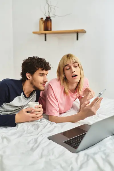 Dos hombres comparten un momento alegre juntos, relajándose en la cama e interactuando con su computadora portátil y teléfono. - foto de stock