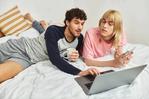 Deux hommes se détendent sur leur lit, l'un tapant sur un ordinateur portable tandis que l'autre s'engage à proximité. — Photo de stock