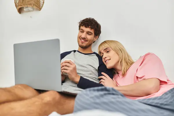 Deux hommes se blottissent sur un lit, profitant d'une boisson chaude et d'un spectacle sur un ordinateur portable. — Photo de stock