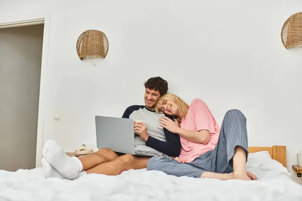 Two men relax on a bed, sharing laughter and warmth while browsing their laptop in a cozy room. — Stock Photo