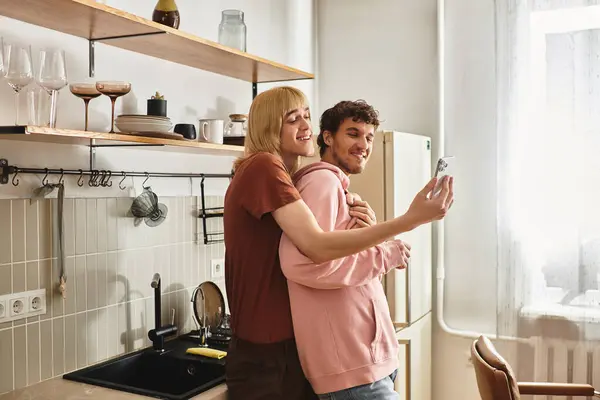 Zwei Männer umarmen sich herzlich in ihrer hellen Küche, während sie einen freudigen Moment mit ihrem Handy festhalten. — Stockfoto