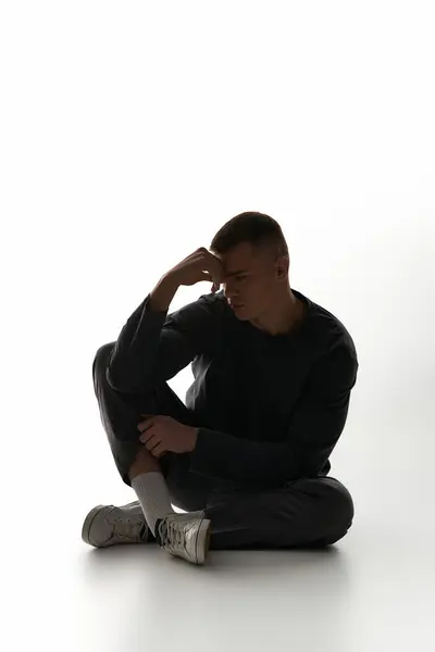 A young man sits pensively, hand on forehead, lost in thought in a minimalistic space. — Stock Photo