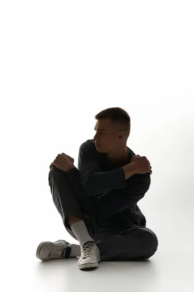 Un jeune homme s'assoit gracieusement avec les bras autour des genoux, perdu dans la pensée sur un fond frais. — Photo de stock