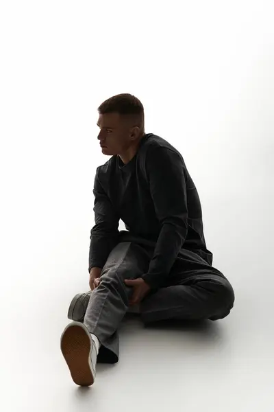 A young man sits on the floor, thoughtfully holding his leg in soft light. — Stock Photo