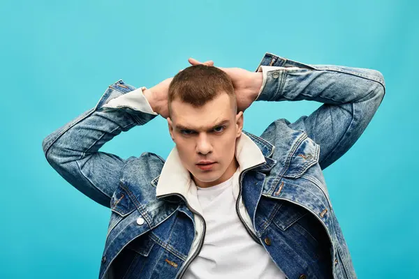 Ein selbstbewusster junger Mann in Jeansjacke posiert verspielt vor hellblauem Hintergrund. — Stockfoto