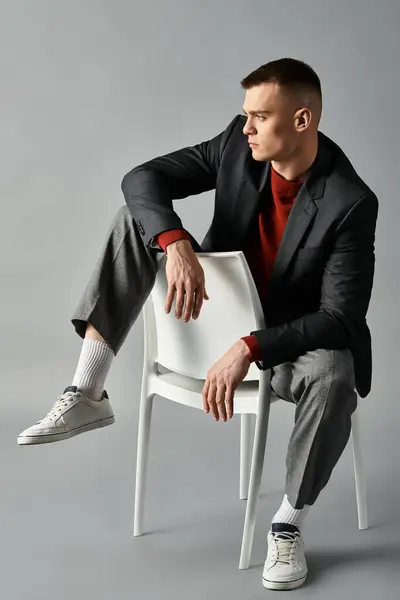 A young man in a stylish suit and sneakers poses dynamically with a chair on a neutral backdrop. — Stock Photo