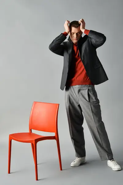 A young man stands thoughtfully beside a vibrant red chair, showcasing his fashionable style. — Stock Photo