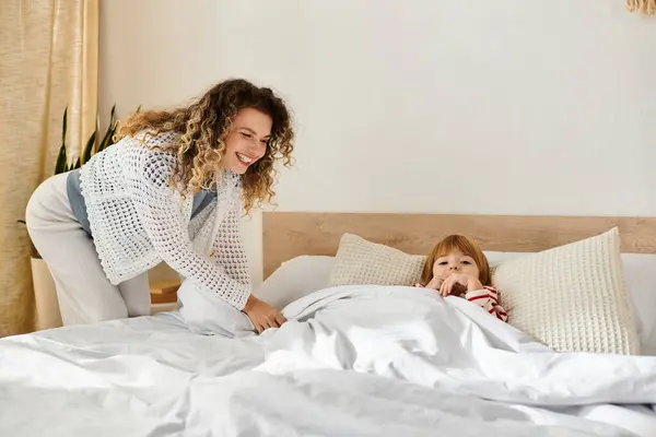 Une mère aide joyeusement sa fille à se blottir dans son lit, montrant leur lien chaleureux à la maison. — Photo de stock