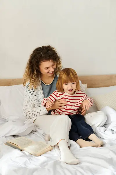 A curly-haired mother enjoys playful time with her daughter on a comfy bed in their modern home. — Stock Photo