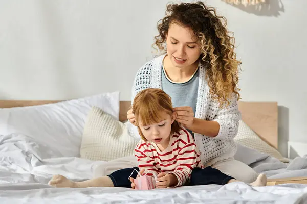 Mother lovingly styles her daughter hair while they relax on the bed in their inviting room. — Stock Photo