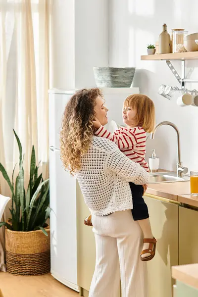 A loving mother holds her curly-haired daughter in a bright kitchen filled with warmth and joy. — Stock Photo