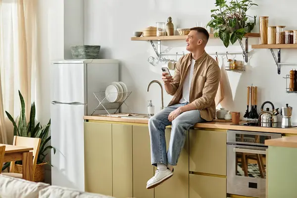 A young man enjoys his time at home, casually sitting on the counter and using his phone. — Stock Photo