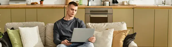 A handsome young man relaxes on a cozy sofa while using his laptop in a modern apartment, banner — Stock Photo