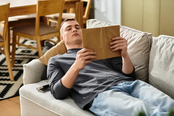 A young man lounges on a comfy couch, engaged with an interesting reading material at home. — Stock Photo
