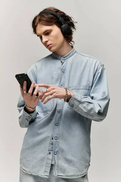 A handsome young man dressed in denim stands focused, listening to music and exploring his phone. — Stock Photo