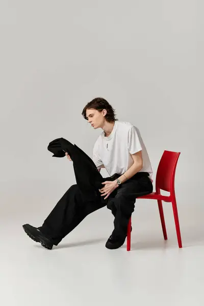A handsome young man in casual attire sits on a vibrant red chair, inspecting a black garment. — Stock Photo