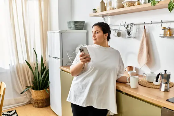 A young plus sized woman stands in a modern kitchen, thoughtfully holding her smartphone. — Stock Photo