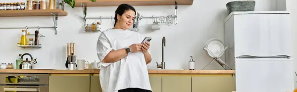 A beautiful plus sized woman stands in her stylish kitchen, happily texting on her smartphone. — Stock Photo