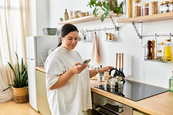 En una cocina elegante, una hermosa mujer joven de tamaño grande sonríe mientras usa su teléfono - foto de stock