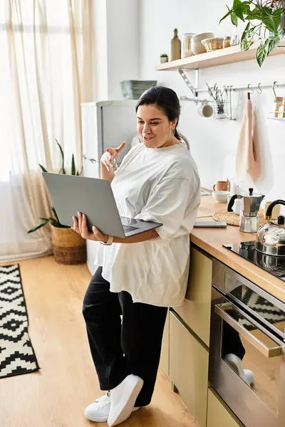 Cucina illuminata dove una donna plus size parla animatamente durante l'utilizzo del suo computer portatile. — Foto stock