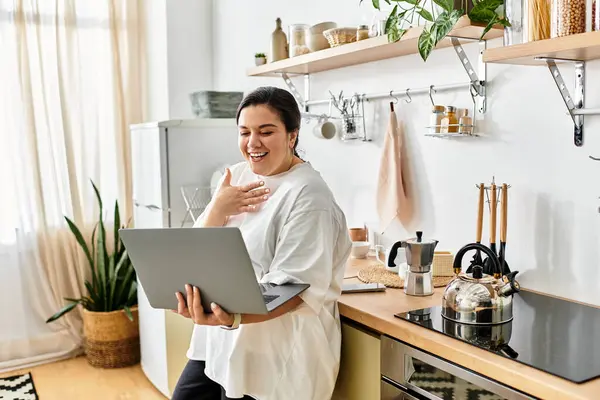 Uma jovem mulher de tamanho grande sorri ao usar seu laptop em um espaço de cozinha brilhante e convidativo. — Fotografia de Stock