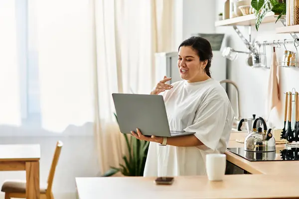 Engaged in conversation, a beautiful young woman stands in her modern kitchen, laptop in hand. — Stock Photo