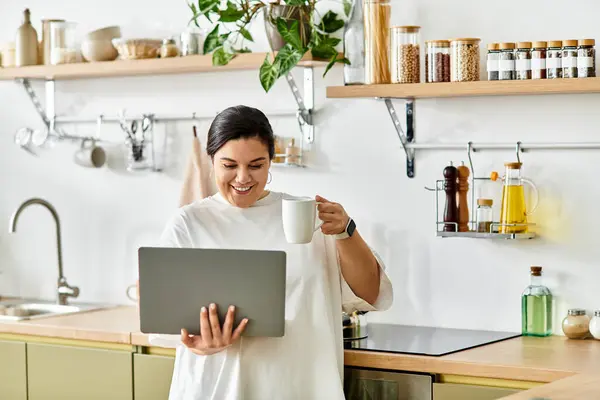 Cucina luminosa con una giovane allegra plus size donna sorseggiando caffè e utilizzando il suo computer portatile. — Foto stock