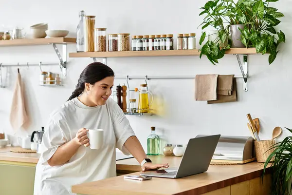 In a cozy kitchen, a young plus sized woman smiles while working on her laptop and sipping coffee. — Stock Photo