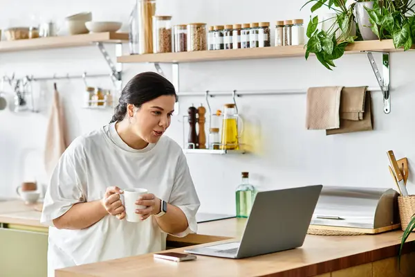 Dans une cuisine élégante, une jeune femme de taille plus sourit avec une boisson chaude, bavardant en ligne. — Photo de stock