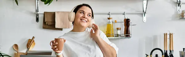 Eine junge Plus-Size-Frau mit freudigem Lächeln hält eine Tasse in schickem Küchenambiente, verloren in der Musik. — Stockfoto