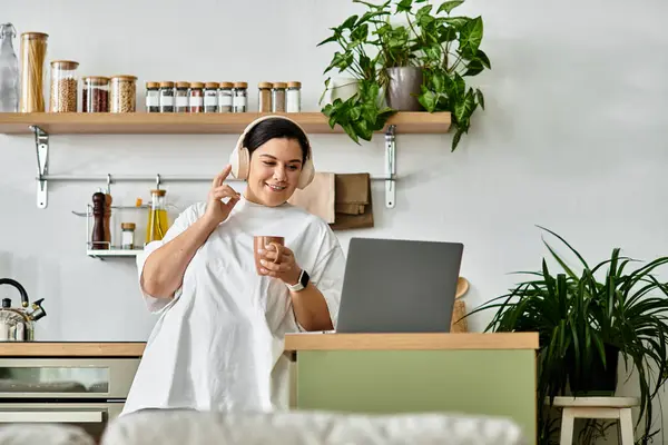 A cheerful plus sized woman with headphones engages with her laptop in a bright kitchen. — Stock Photo