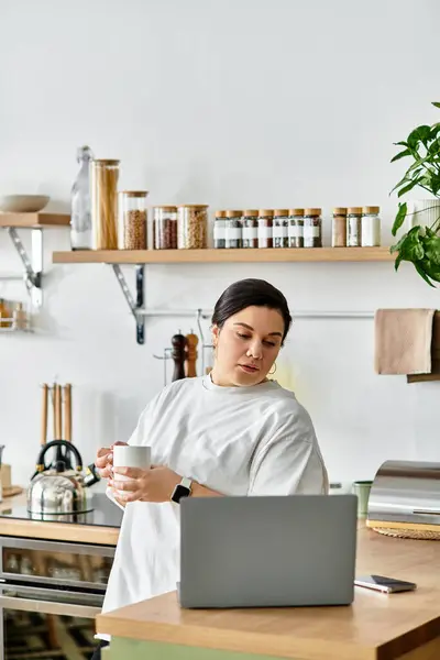 A young woman relaxes in a modern kitchen, holding a cup and focused on her laptop. — Stock Photo
