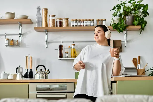 A cheerful young woman relaxes in her stylish kitchen, holding a drink while wearing headphones. — Stock Photo
