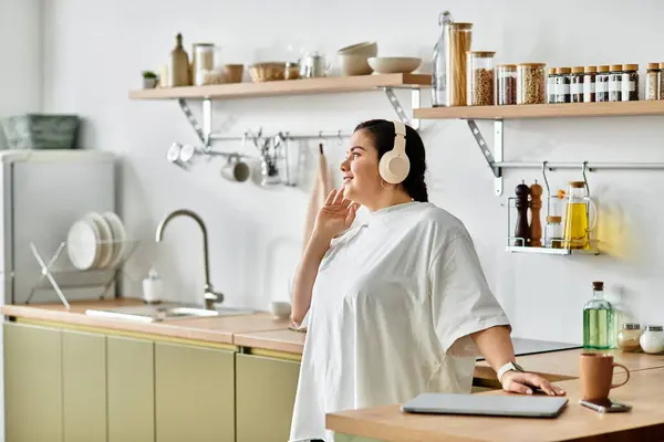 Plus taille femme porte des écouteurs et des sourires tout en se tenant dans un espace de cuisine chic. — Photo de stock