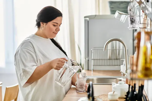 Una joven mujer de tamaño grande vierte agua de una jarra de vidrio en un vaso de cristal en casa, sonriendo. - foto de stock