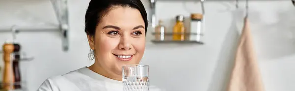 Sorridente giovane donna plus size tiene un bicchiere d'acqua in una cucina ben arredata con calore. — Foto stock