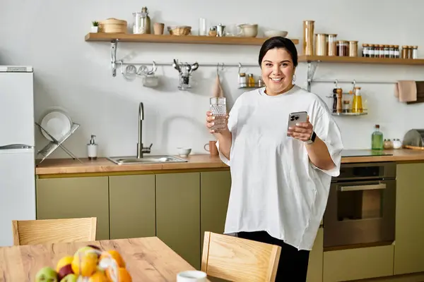 Smiling plus sized woman holds a drink and phone while standing in a stylish kitchen space. — Stock Photo