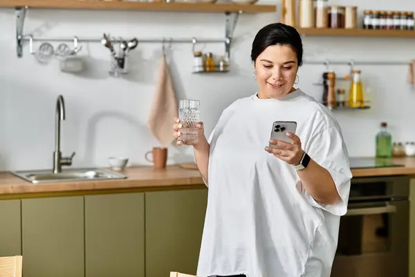 Une jeune femme joyeuse tient un verre d'eau en regardant son téléphone dans une cuisine moderne. — Photo de stock