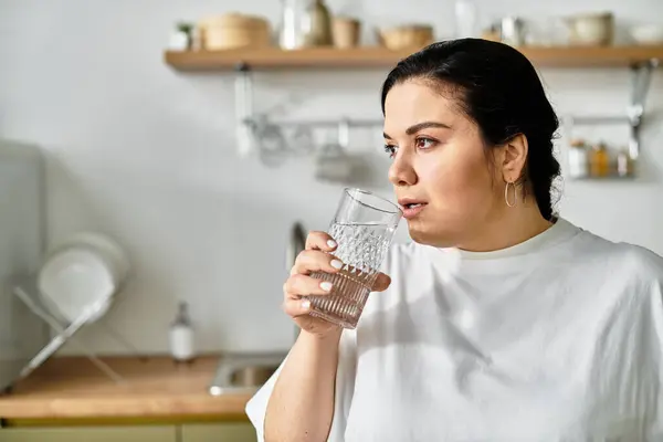 Una encantadora mujer joven de tamaño grande se relaja mientras bebe agua en una elegante área de cocina. - foto de stock