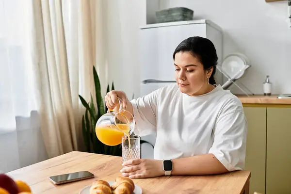 Una joven mujer de tamaño grande vierte jugo de naranja en un vaso en una mesa de madera, saboreando su mañana. - foto de stock