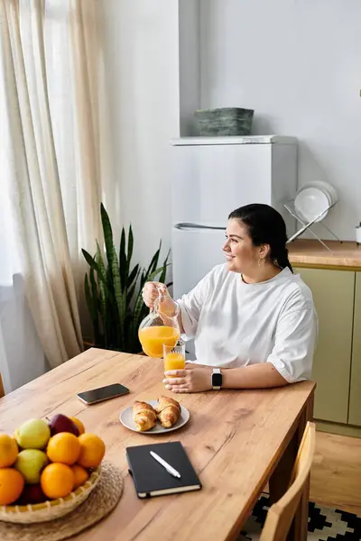 Una joven, hermosa mujer de tamaño grande vierte jugo de naranja en una mesa de madera adornada con frutas. - foto de stock