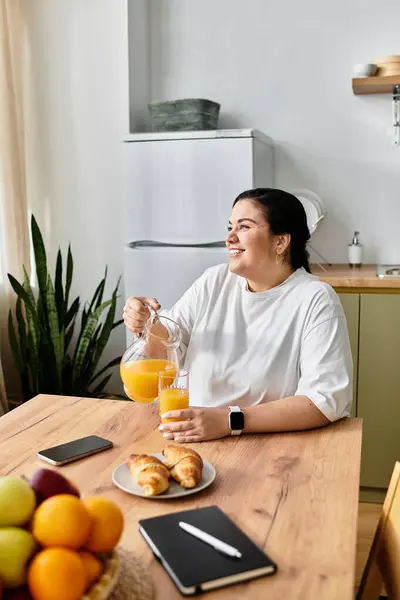 Eine junge Plus-Size-Frau schenkt Orangensaft ein, während sie an einem gemütlichen Küchentisch lächelt. — Stockfoto