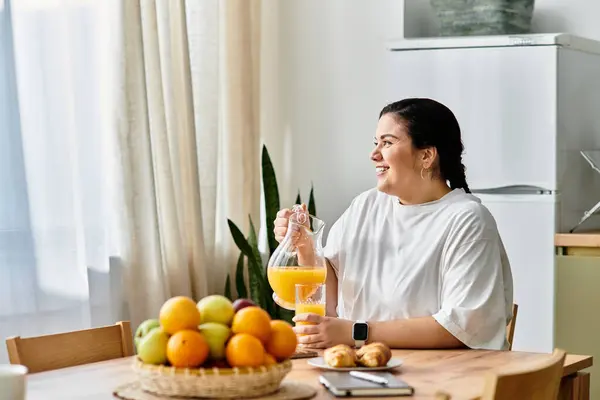 Una joven mujer de tamaño grande sonríe mientras vierte jugo de naranja en un vaso, rodeado de fruta fresca. - foto de stock