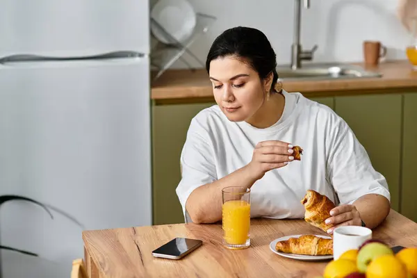 A young plus sized woman savors pastries and juice while checking her phone in a cozy kitchen. — Stock Photo