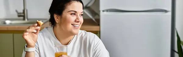 A young plus sized woman smiles while enjoying a snack and drink in her cozy kitchen. — Stock Photo